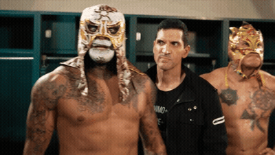 A Man In A Black Shirt Stands Next To A Man Wearing A Mask GIF