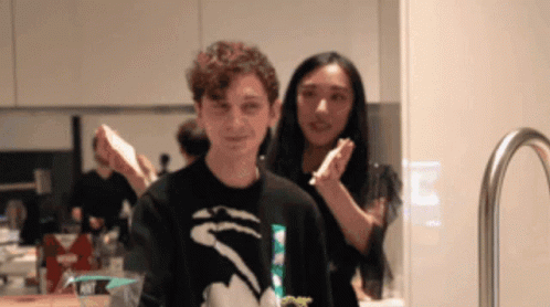 A Man In A Black Shirt With A Shark On It Stands Next To A Woman In A Kitchen GIF