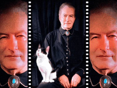 A Man In A Black Shirt With A Turquoise Pendant Sits Next To A White Cat GIF