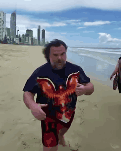 A Man In A Black Shirt With An Eagle On It Is Walking On The Beach GIF