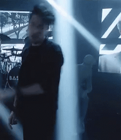 A Man In A Black Shirt With The Word Bas On The Back Is Standing In A Dark Room . GIF