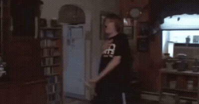 A Man In A Black Shirt With The Word Cats On It Is Standing In A Living Room GIF