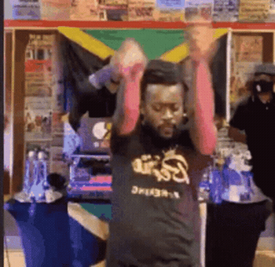 A Man In A Black Shirt With The Word Reggae On It Is Dancing In Front Of A Jamaican Flag . GIF