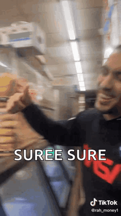 A Man In A Black Shirt With The Word Suree Sure On It Is Standing In Front Of A Refrigerator In A Store . GIF