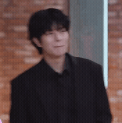A Man In A Black Suit And Black Shirt Is Making A Funny Face While Standing In Front Of A Brick Wall . GIF