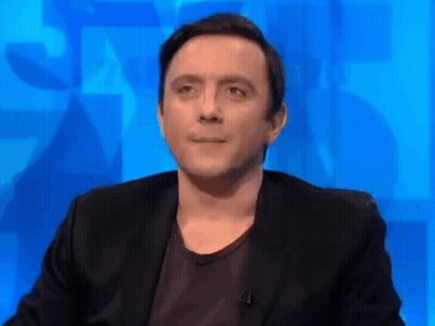 A Man In A Black Suit And Black Shirt Is Sitting In Front Of A Blue Wall . GIF