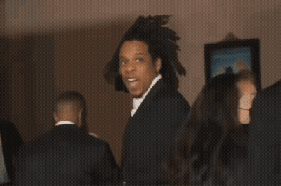 A Man In A Black Suit And White Tie Is Standing In A Room GIF