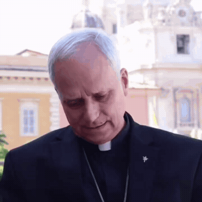 A Man In A Black Suit Has A Badge On His Lapel That Says ' Cardinal ' On It GIF