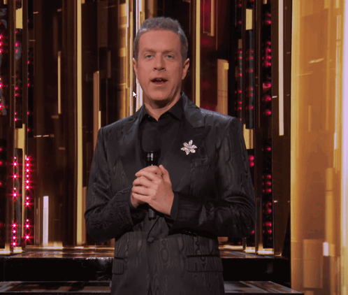 A Man In A Black Suit Holds A Microphone In Front Of A Gold Wall GIF
