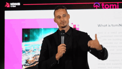 A Man In A Black Suit Holds A Microphone In Front Of A Screen That Says Tomi GIF