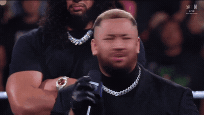 A Man In A Black Suit Is Holding A Microphone In Front Of A Crowd That Is Watching A Live Wrestling Event GIF