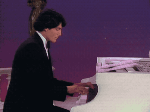 A Man In A Black Suit Is Playing A White Grand Piano GIF