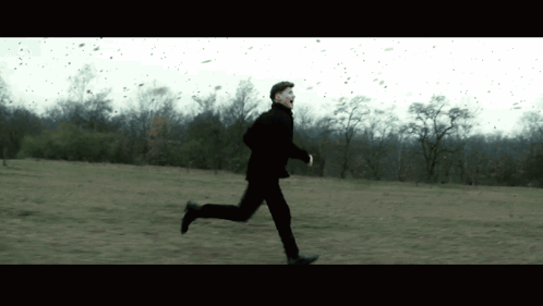 A Man In A Black Suit Is Running Through A Field GIF