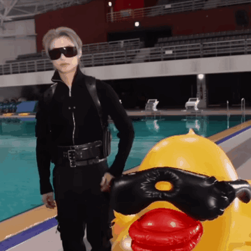 A Man In A Black Suit Is Standing Next To A Yellow Rubber Duck GIF