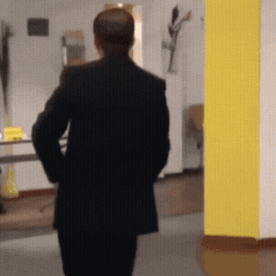 A Man In A Black Suit Is Walking In A Hallway With His Hands In His Pockets GIF
