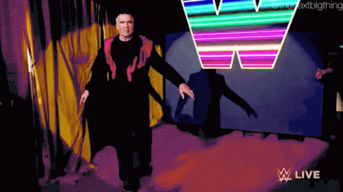 A Man In A Black Suit Is Walking In Front Of A Neon W Sign GIF