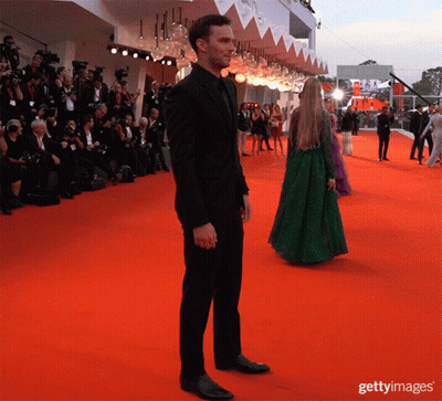 A Man In A Black Suit Stands On A Red Carpet With A Woman In A Green Dress Standing Behind Him GIF