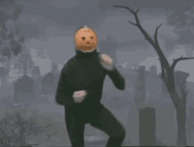 A Man In A Black Suit With A Pumpkin On His Head Is Standing In A Cemetery . GIF