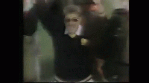 A Man In A Black Sweater And Sunglasses Is Dancing In Front Of A Crowd Of People . GIF