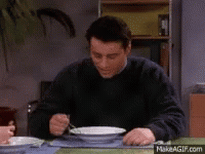 A Man In A Black Sweater Is Sitting At A Table Eating From A Bowl With A Fork . GIF
