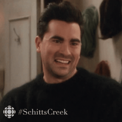 A Man In A Black Sweater Is Smiling In A Room With A Sign That Says Schitts Creek . GIF