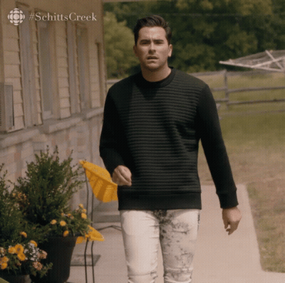 A Man In A Black Sweater Is Walking Down A Sidewalk In Front Of A House With #Schittscreek On The Bottom GIF