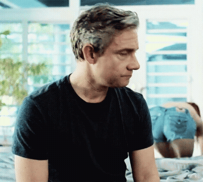 A Man In A Black T-Shirt Is Sitting On A Bed With His Eyes Closed GIF