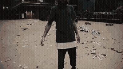 A Man In A Black T-Shirt Is Standing In A Dirt Area GIF