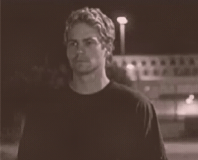 A Man In A Black T-Shirt Is Standing In Front Of A Building At Night . GIF
