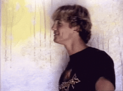 A Man In A Black T-Shirt Is Standing In Front Of A Wall And Smiling . GIF