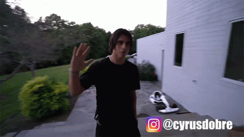 A Man In A Black T-Shirt Is Standing In Front Of A White Building With The Hashtag @Cyrusdobre On The Bottom GIF