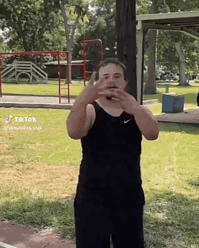 A Man In A Black Tank Top And Black Shorts Is Standing In A Park With His Hands On His Face . GIF