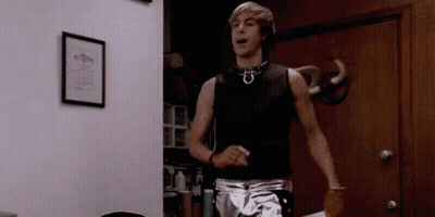 A Man In A Black Tank Top And Silver Shorts Is Standing In Front Of A Door . GIF