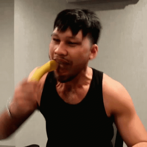 A Man In A Black Tank Top Is Biting A Banana GIF