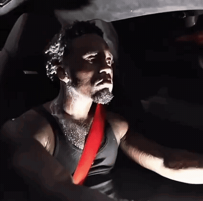 A Man In A Black Tank Top Is Driving A Car With A Red Seat Belt GIF