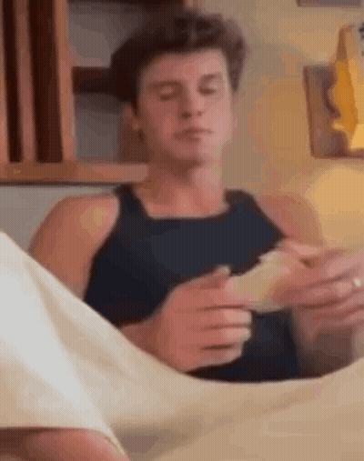 A Man In A Black Tank Top Is Sitting On A Bed Holding A Piece Of Paper . GIF