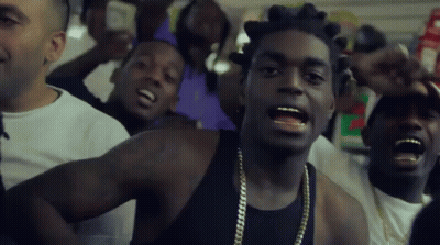 A Man In A Black Tank Top With Gold Chains Around His Neck Is Surrounded By Other Men GIF