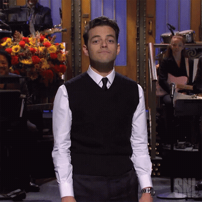 A Man In A Black Vest And White Shirt Stands In Front Of A Snl Sign GIF