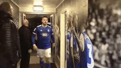 A Man In A Blue And White Soccer Uniform Is Walking Down A Hallway With Two Men . GIF