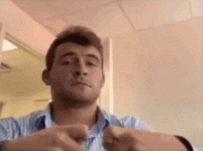 A Man In A Blue And White Striped Shirt Is Making A Funny Face While Holding His Hands Together . GIF