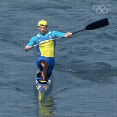 A Man In A Blue And Yellow Outfit Is Paddling A Canoe In The Water With The Olympic Rings In The Background GIF