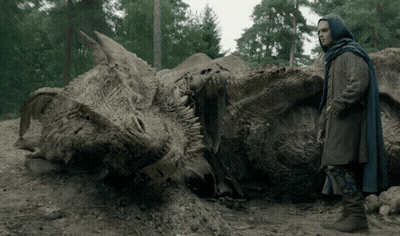 A Man In A Blue Cape Stands Next To A Large Dragon Skeleton GIF