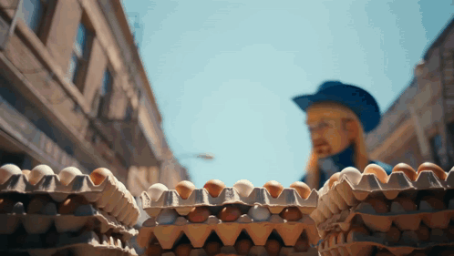 A Man In A Blue Hat Is Standing Behind A Stack Of Eggs GIF