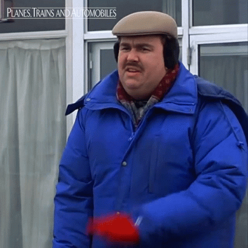 A Man In A Blue Jacket And Hat Is Standing In Front Of A Window With Planes Trains And Automobiles Written Above Him GIF