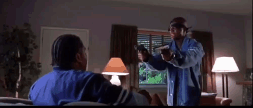 A Man In A Blue Jacket Is Pointing A Gun At Another Man Sitting On A Couch . GIF