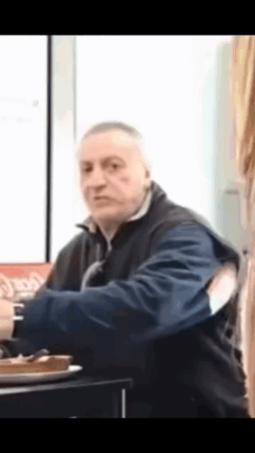A Man In A Blue Jacket Is Sitting At A Table With A Plate Of Food . GIF