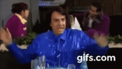 A Man In A Blue Jacket Is Sitting At A Table With His Arms Outstretched In Front Of A Group Of People . GIF