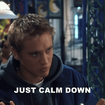 A Man In A Blue Jacket Is Sitting At A Table With The Words `` Just Calm Down '' Written On The Screen . GIF
