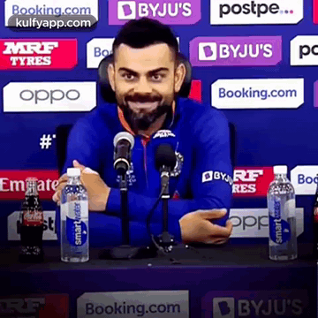 A Man In A Blue Jacket Is Sitting In Front Of A Microphone Holding A Bottle Of Water . GIF