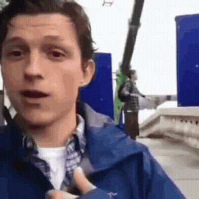 A Man In A Blue Jacket Is Standing In Front Of A Roller Coaster And Pointing At It . GIF